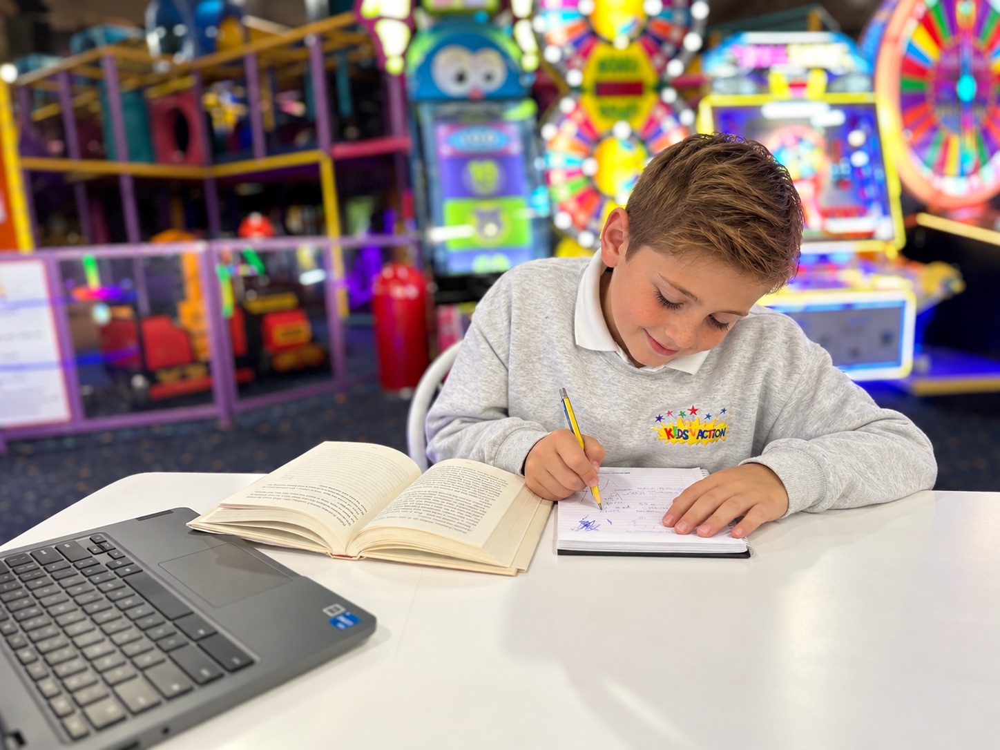 A boy doing homework at a table in Kids 'N Action, with the play area and arcade behind him